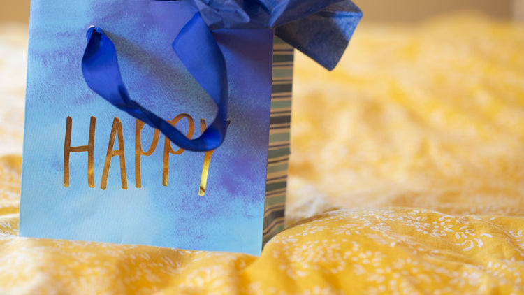 Blaue Geschenktüte mit Aufschrift Happy auf gelbem Tuch – Geschenkidee für Selbstfürsorge und kleine Auszeiten